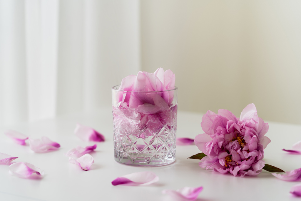 πολύπλευρο γυαλί με τονωτικό και πέταλα κοντά σε ροζ παιώνια σε λευκό τραπέζι και γκρι φόντο - Φωτογραφία, εικόνα