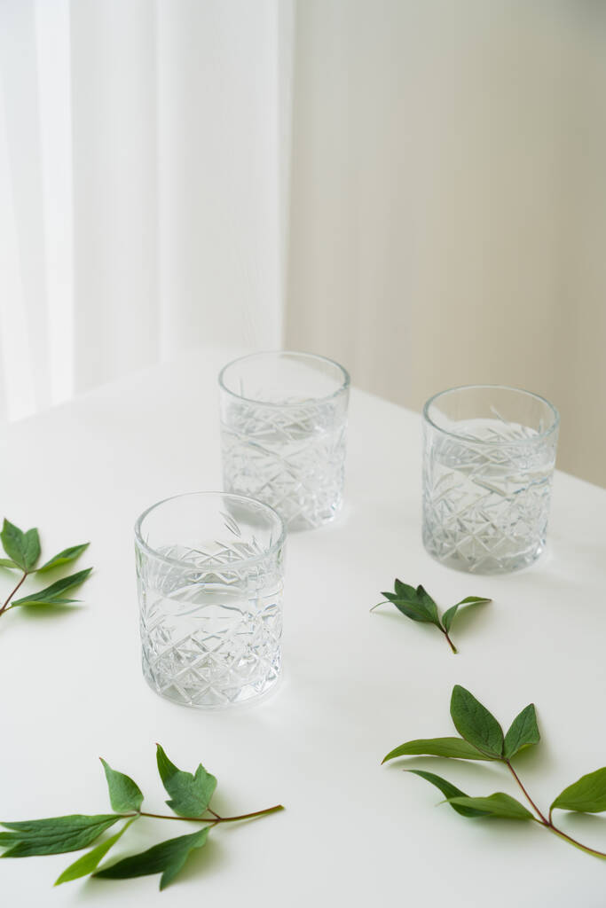 zelené listy a křišťálové sklenice s čistou vodou na bílém stolku a šedé pozadí - Fotografie, Obrázek