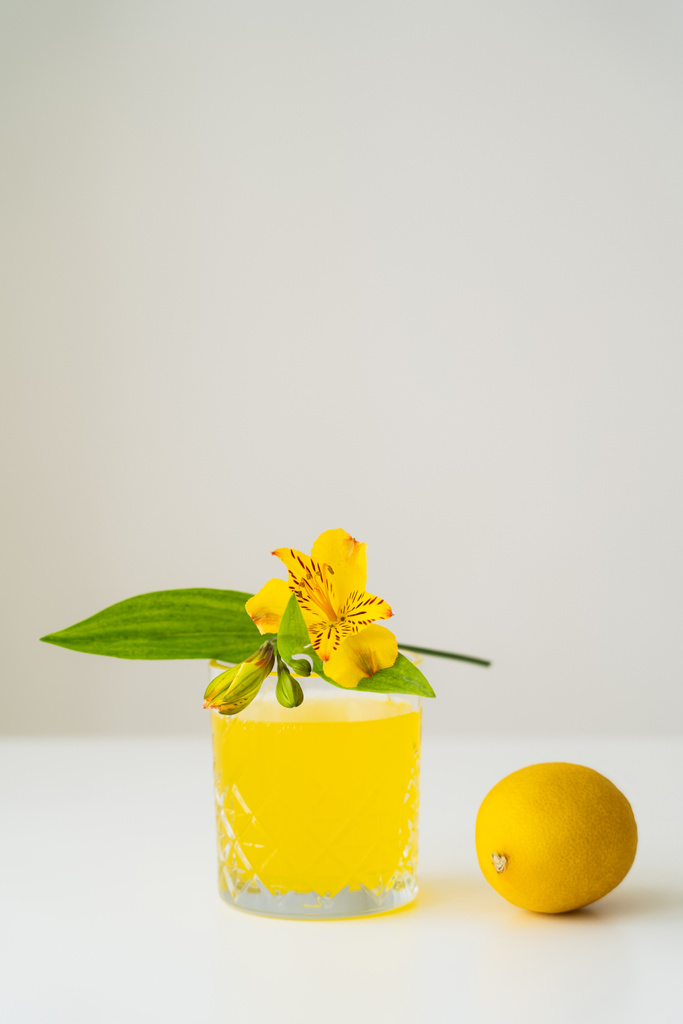 yellow peruvian lily and whole lemon near glass of citrus tonic on white surface isolated on grey - Photo, Image