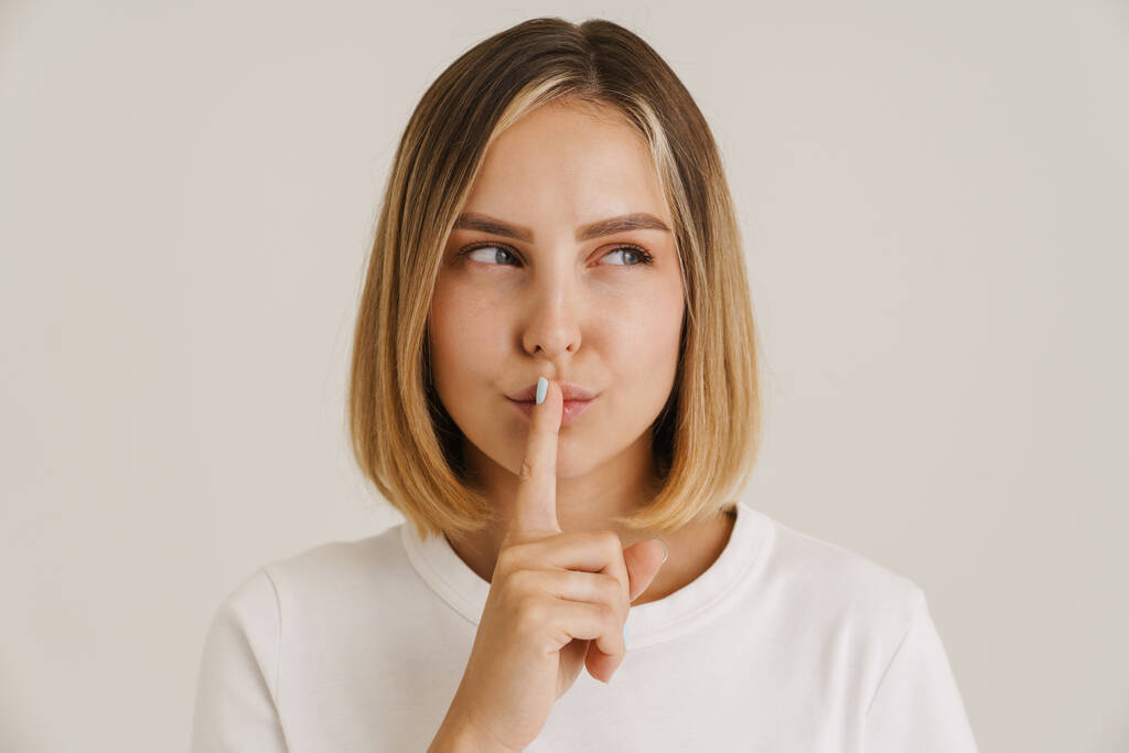 Young blonde woman looking aside while showing silence gesture isolated over white background - Photo, Image