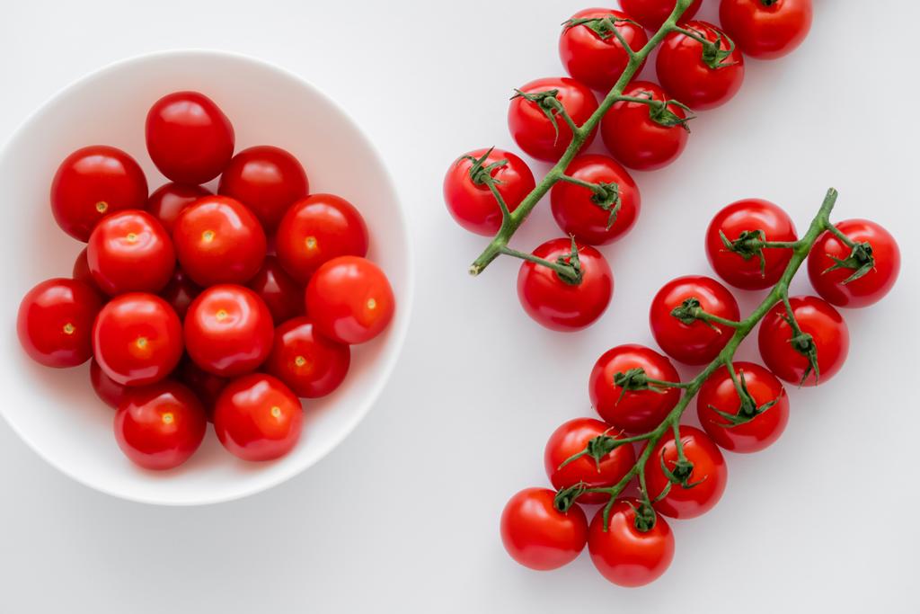 Vista superior de tomates cherry rojos maduros en ramas y en tazón sobre fondo blanco  - Foto, Imagen