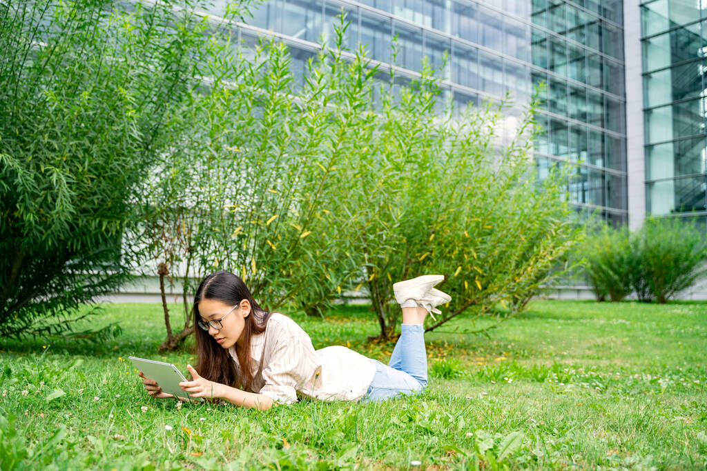 Jeune femme asiatique heureuse thaïlandaise, vietnamienne ou chinoise en vêtements décontractés et lunettes allongées sur l'herbe verte prêt à l'extérieur près de l'université travaillant apprentissage de la lecture ou bavarder à l'aide d'une tablette le week-end - Photo, image