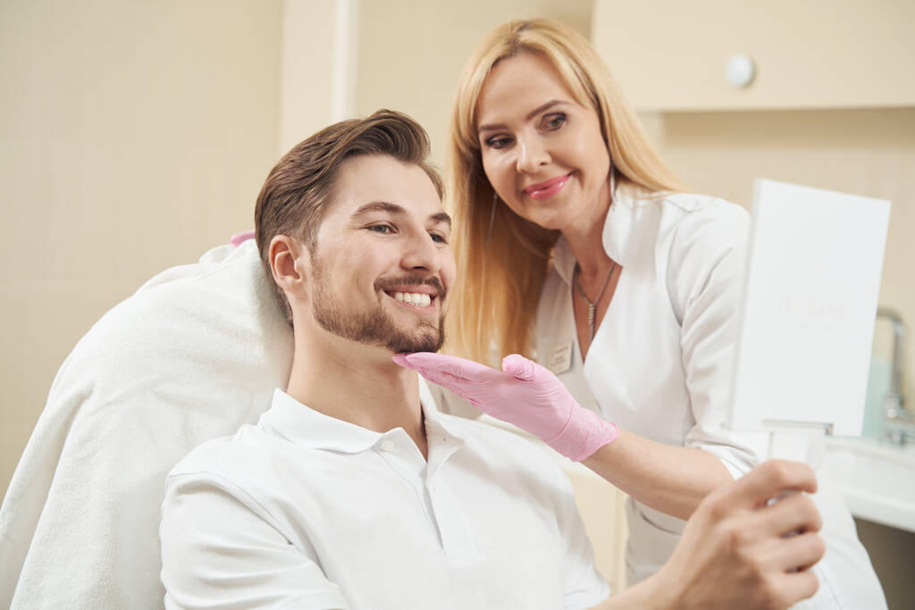 Kosmetolog wspiera mężczyznę przez podbródek podczas gdy on daje uśmiech w lustro ręczne przed nim - Zdjęcie, obraz