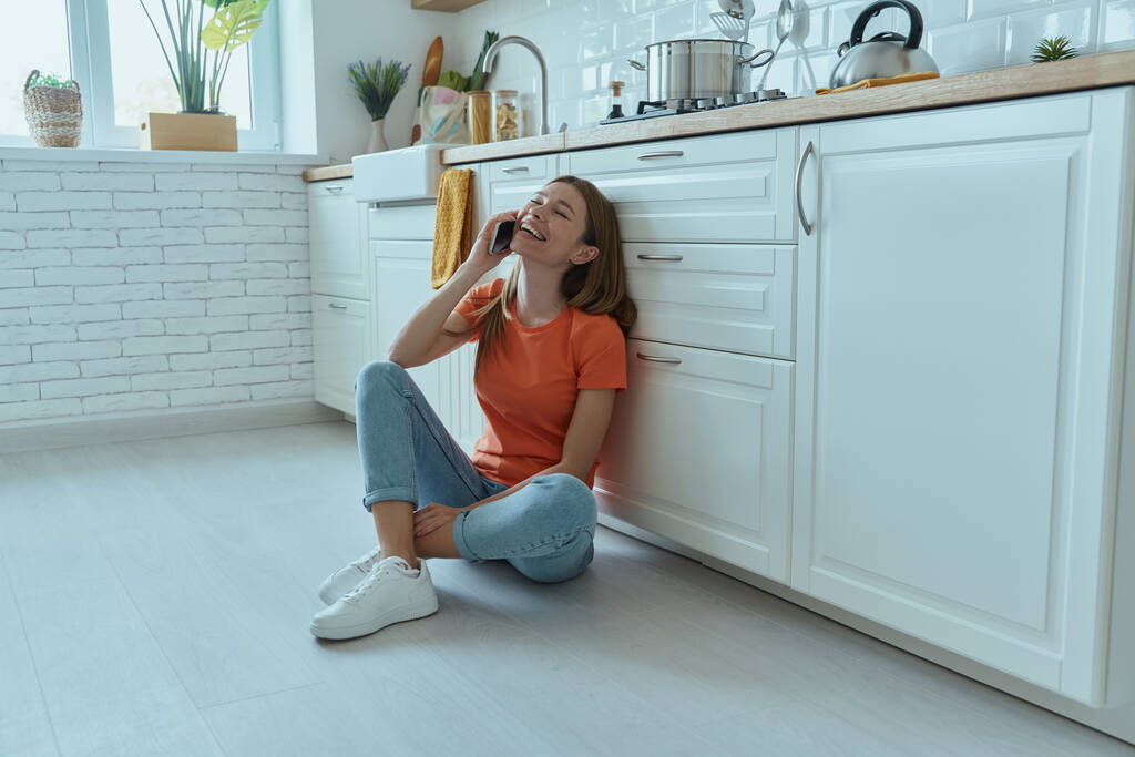 Jovem feliz falando no telefone celular enquanto sentado no chão na cozinha doméstica - Foto, Imagem