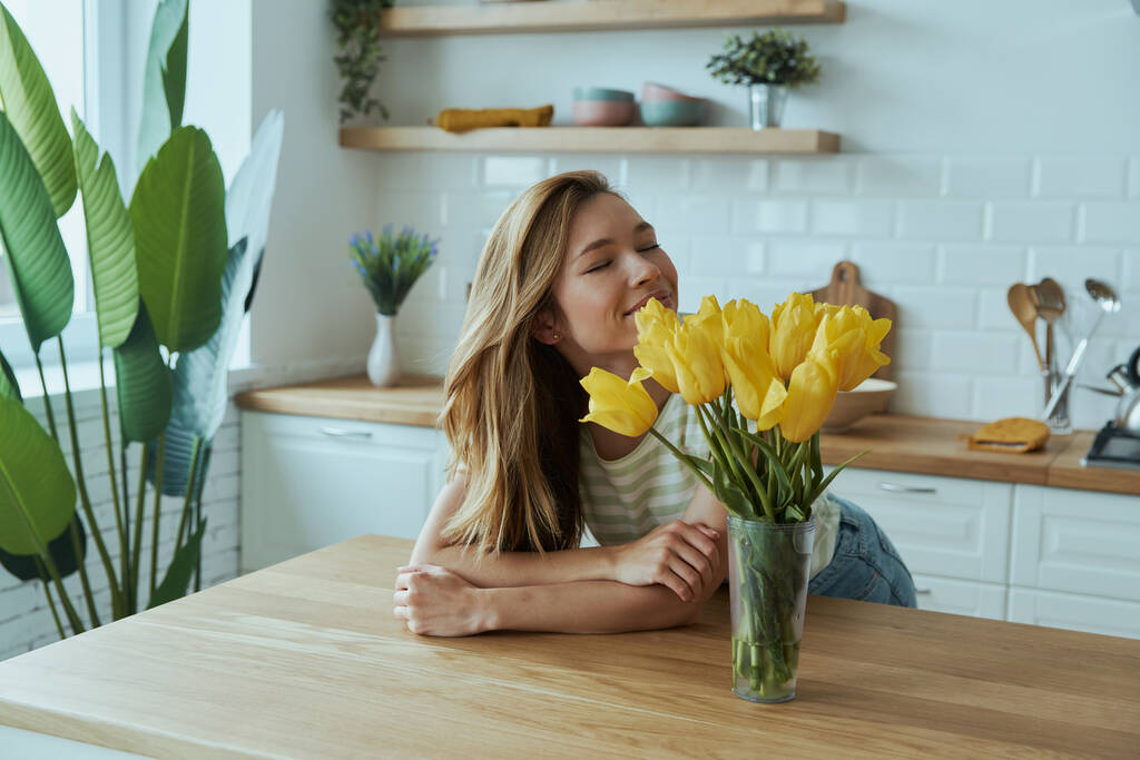 Jovem feliz inclinada na mesa da cozinha e cheirando tulipas amarelas - Foto, Imagem