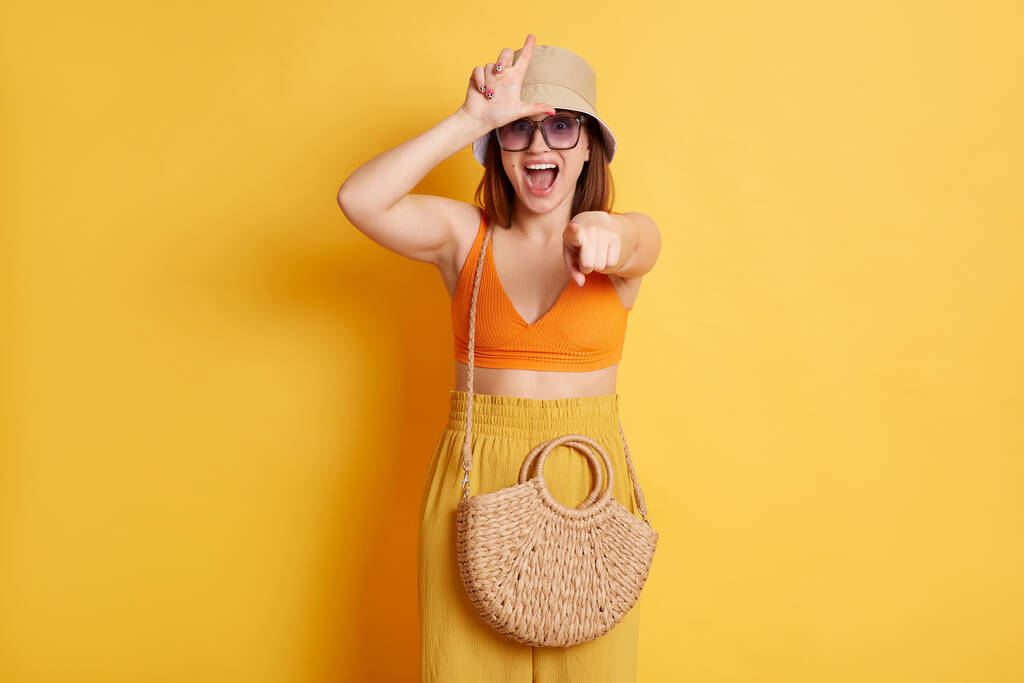 Image d'une belle jeune femme excitée portant un maillot de bain, un short et un panama, posant isolée sur fond jaune, montrant un geste plus lâche et pointant du doigt la caméra. - Photo, image