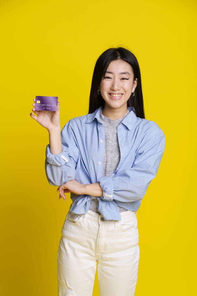 Посміхнена азійська дівчина тримає кредит, дебетова картка в руках. Азійська дівчина в синій сорочці з макетом на жовтому тлі. Концепція E-банкінгу. Концепція електронної комерції. - Фото, зображення