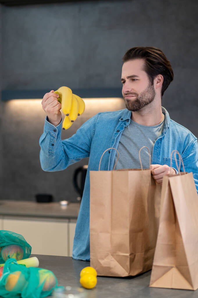 Decepción. Joven barbudo con mueca mirando plátanos en la mano parado cerca de la mesa con compras en la cocina en casa - Foto, imagen