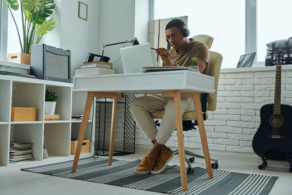 Koncentrált fiatal kevert verseny férfi fülhallgató segítségével laptop, miközben dolgozik az irodában - Fotó, kép