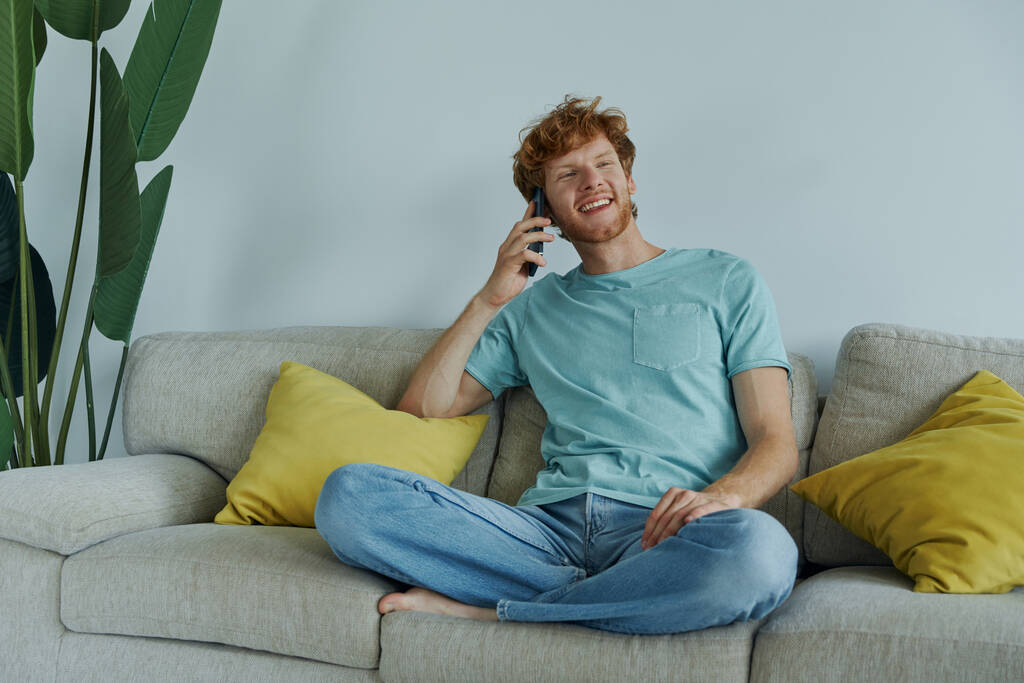 gelukkig roodharige man praten op mobiele telefoon terwijl ontspannen op de bank thuis - Foto, afbeelding