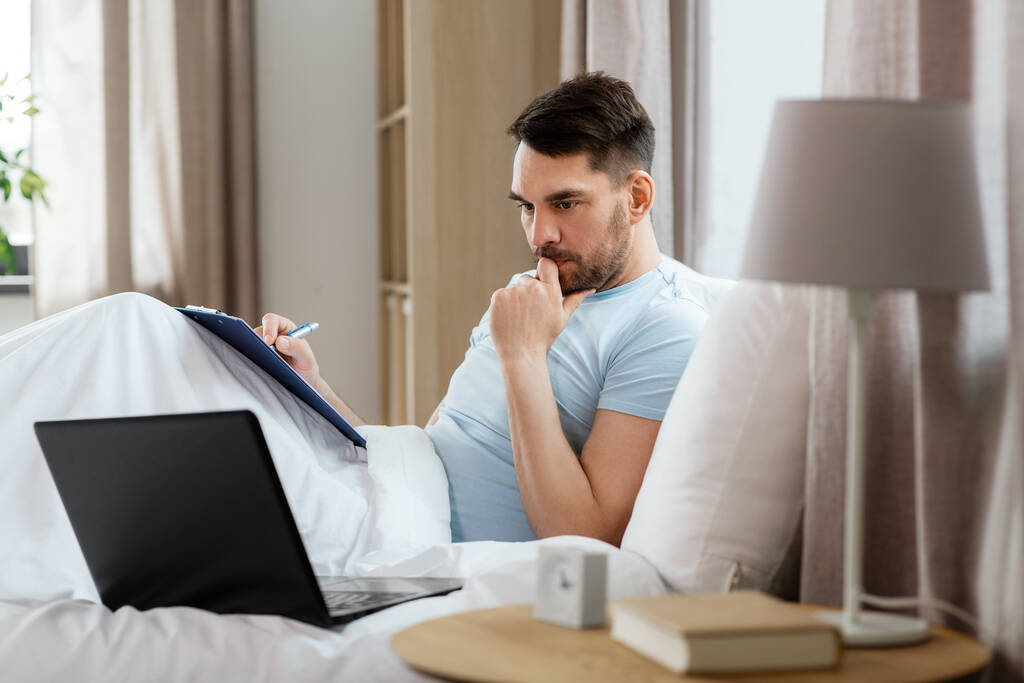 mensen, technologie en remote job concept - man met laptop computer en klembord werken in bed thuis slaapkamer - Foto, afbeelding