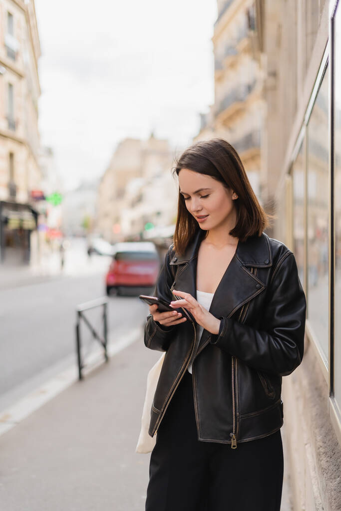 mujer joven en chaqueta de cuero negro mensajería en el teléfono inteligente en la calle en París  - Foto, imagen