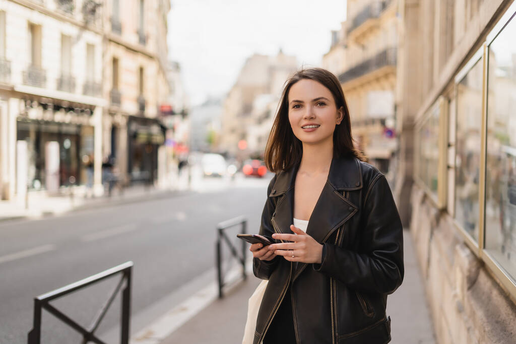 alegre joven mujer en chaqueta de cuero negro celebración de teléfono inteligente en la calle en París  - Foto, Imagen