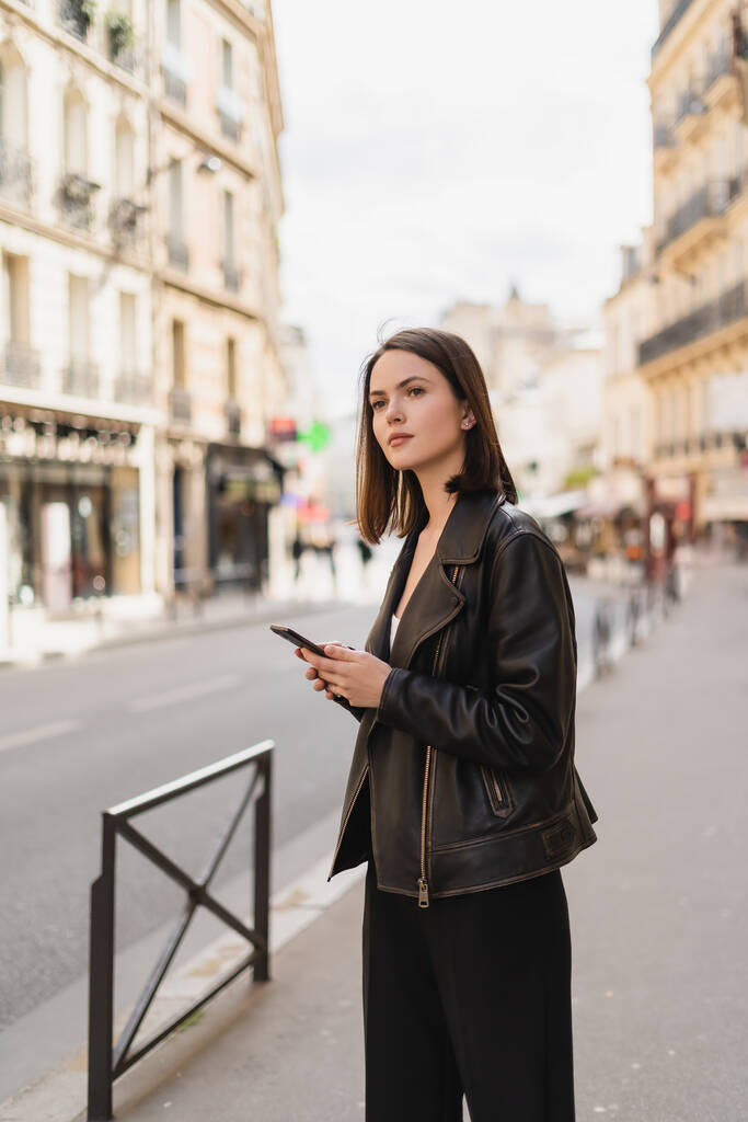 young woman in black leather jacket holding smartphone on street in paris  - Photo, Image