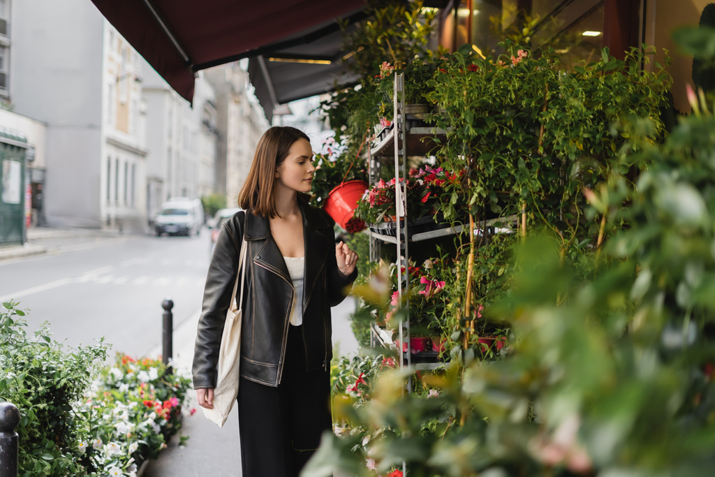 mujer joven y elegante con bolsa de compras de lona mirando plantas en maceta verde en la calle en París  - Foto, imagen