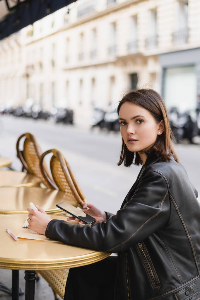 jovem mulher em jaqueta de couro segurando smartphone perto notebook na mesa redonda no terraço do café ao ar livre  - Foto, Imagem