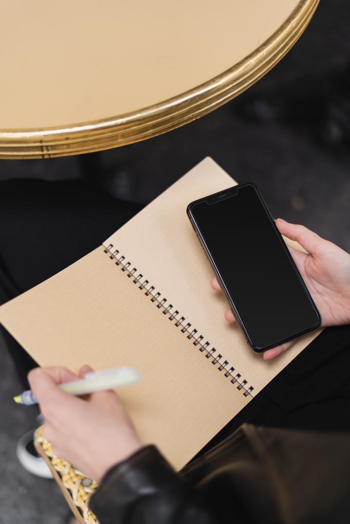 vysoký úhel pohledu ženy držící smartphone s prázdnou obrazovkou a notebook u kulatého stolu - Fotografie, Obrázek