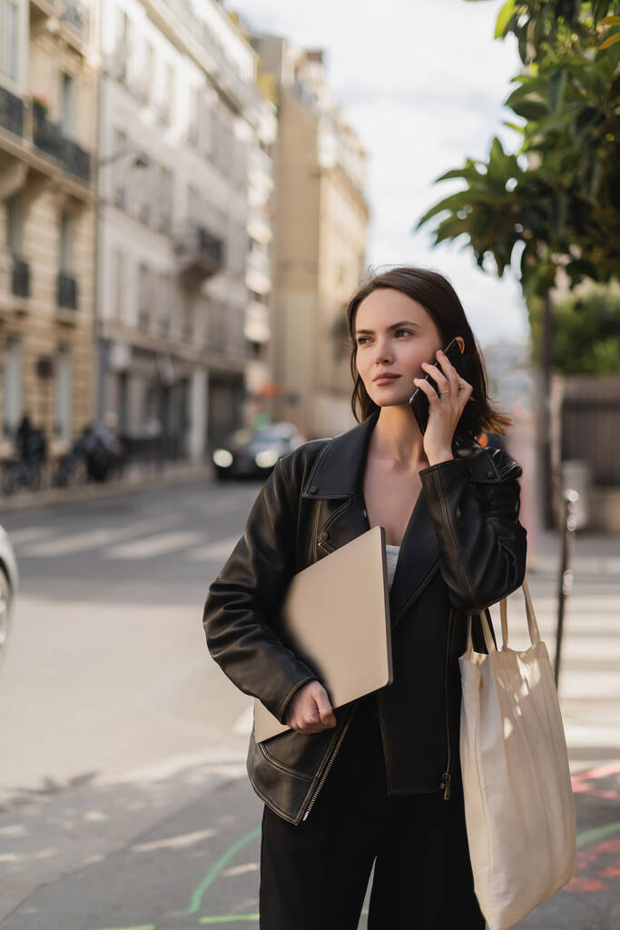 молодой фрилансер в черной кожаной куртке держит ноутбук и разговаривает по смартфону на улице в Париже - Фото, изображение