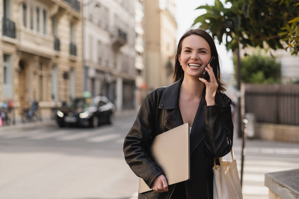 feliz freelancer en chaqueta de cuero negro sosteniendo portátil y hablando en el teléfono inteligente en la calle en París - Foto, imagen