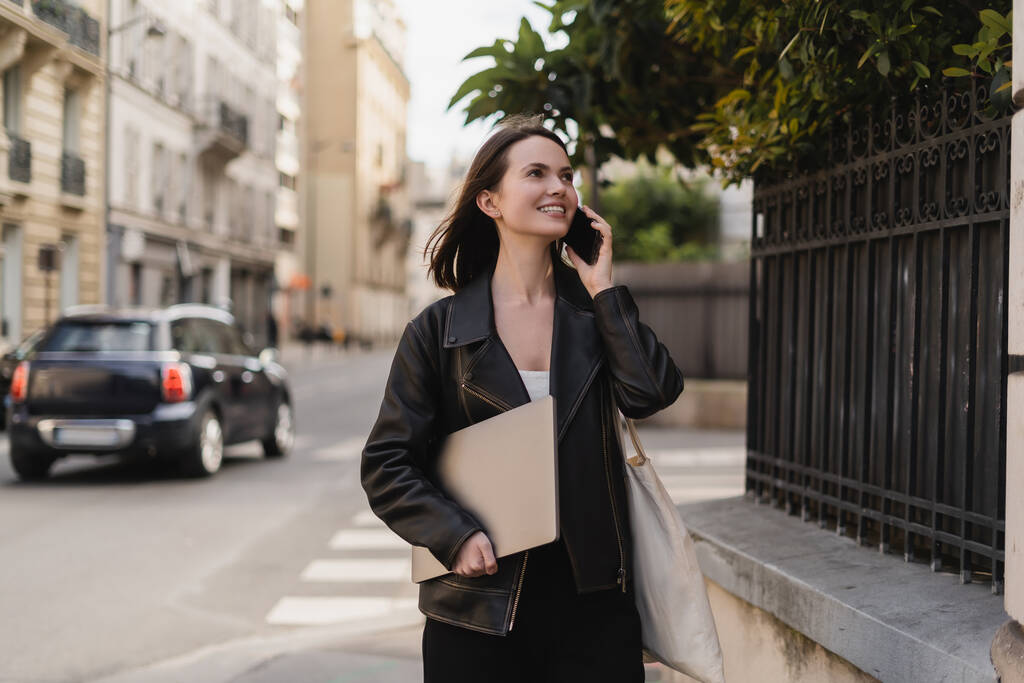 веселый фрилансер в черной кожаной куртке держит ноутбук и разговаривает по смартфону на улице в Париже - Фото, изображение