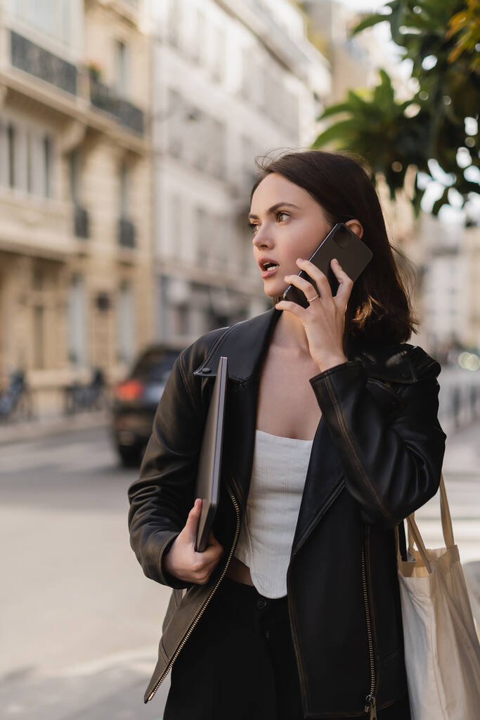 Jeune pigiste veste en cuir noir tenant ordinateur portable et parlant sur téléphone portable dans la rue à Paris - Photo, image