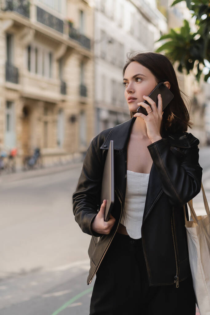 jovem mulher em jaqueta de couro preto segurando laptop e falando no smartphone na rua em paris - Foto, Imagem
