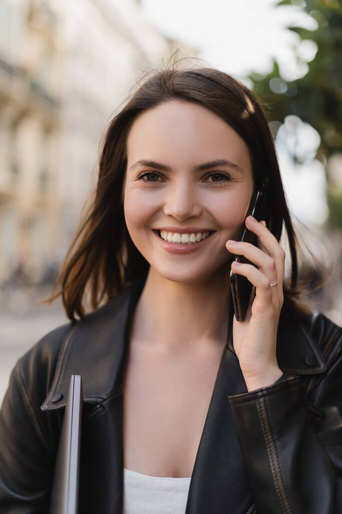 ノートパソコンを持ちパリの路上でスマートフォンで話している黒い革のジャケットの陽気な女性の肖像画 - 写真・画像