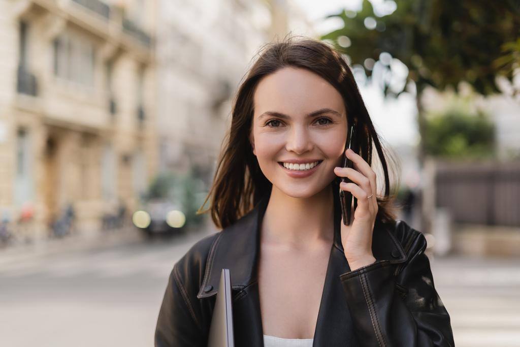 Siyah deri ceketli mutlu bir kadının portresi dizüstü bilgisayar tutuyor ve Paris 'te bir sokakta akıllı telefondan konuşuyor. - Fotoğraf, Görsel