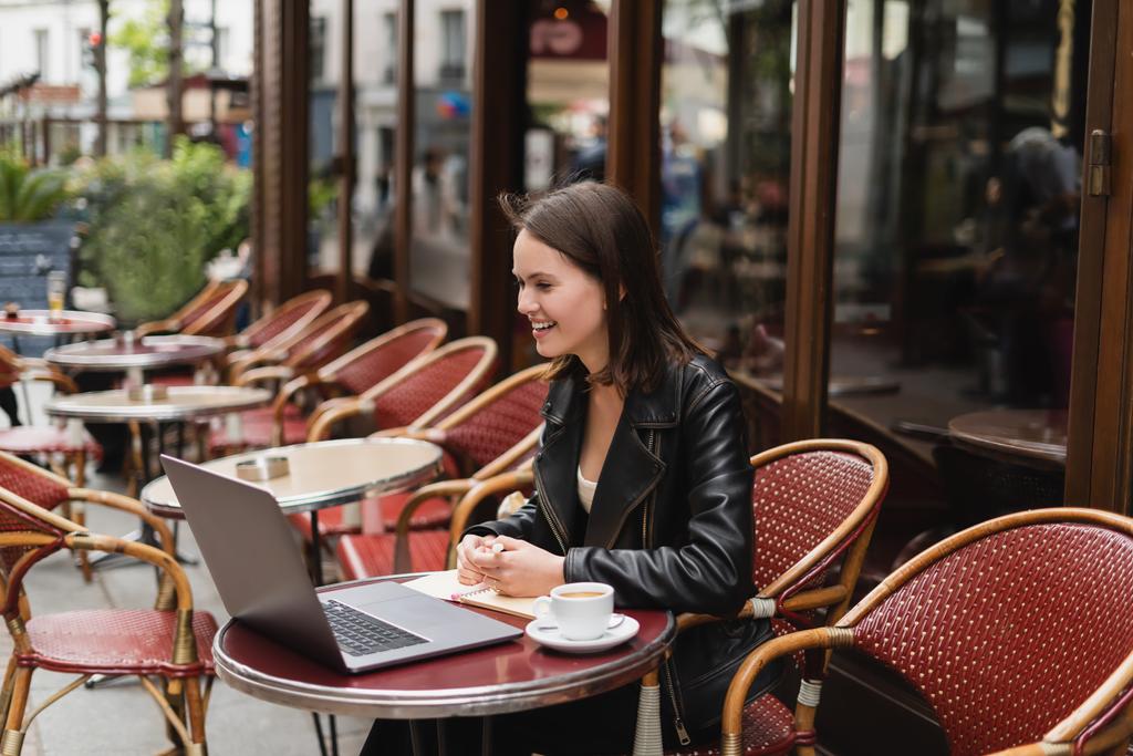 glimlachende freelancer in zwart jasje op zoek naar laptop in de buurt van kopje koffie op tafel in Franse outdoor cafe  - Foto, afbeelding