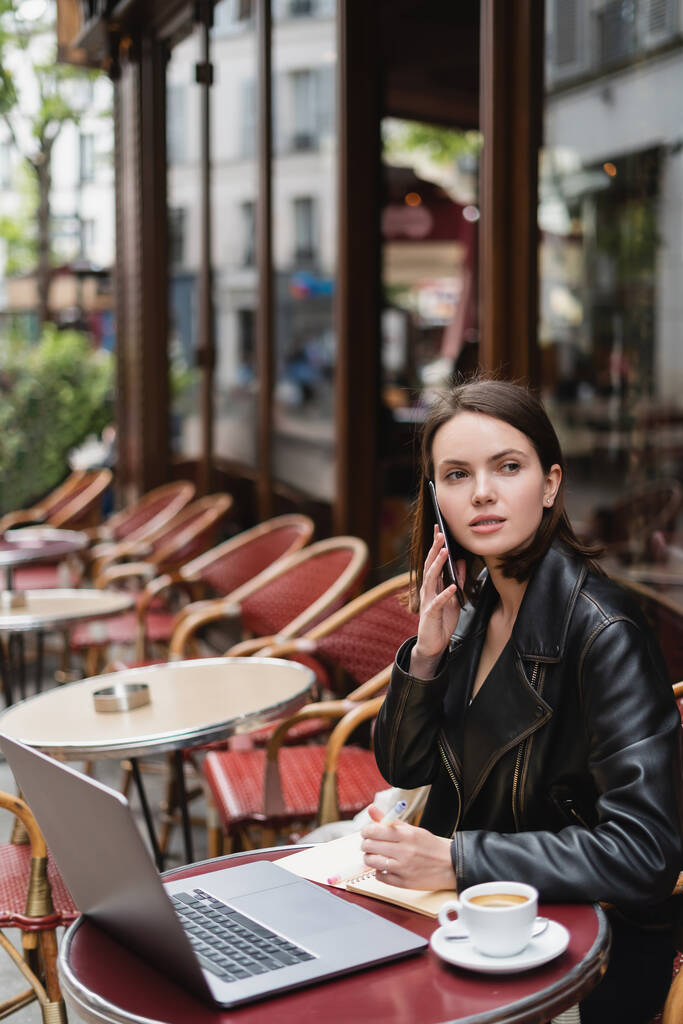 joven freelancer en chaqueta negra hablando en smartphone cerca de portátil y taza de café en la mesa en francés café al aire libre  - Foto, imagen