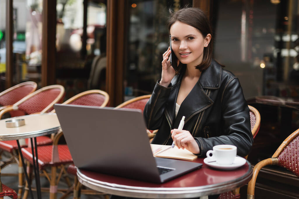 freelance souriant en veste noire parlant sur smartphone près d'un ordinateur portable et tasse de café sur la table dans un café extérieur à Paris - Photo, image
