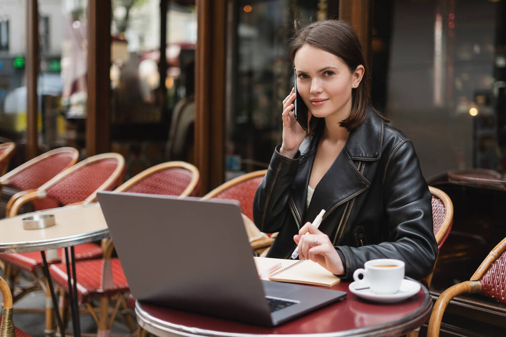 junge Freiberuflerin in schwarzer Jacke unterhält sich auf dem Smartphone neben Laptop und einer Tasse Kaffee auf dem Tisch in einem Café in Paris - Foto, Bild