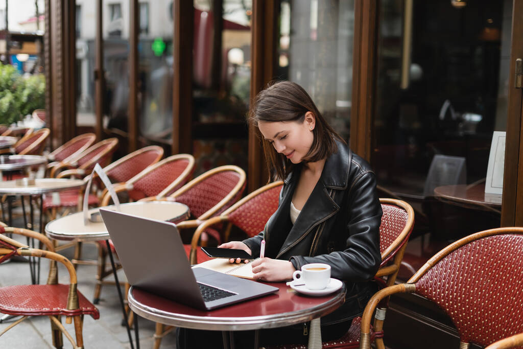 молодой фрилансер в черной куртке, держащий смартфон и пишущий возле ноутбука и чашку кофе во французском открытом кафе  - Фото, изображение