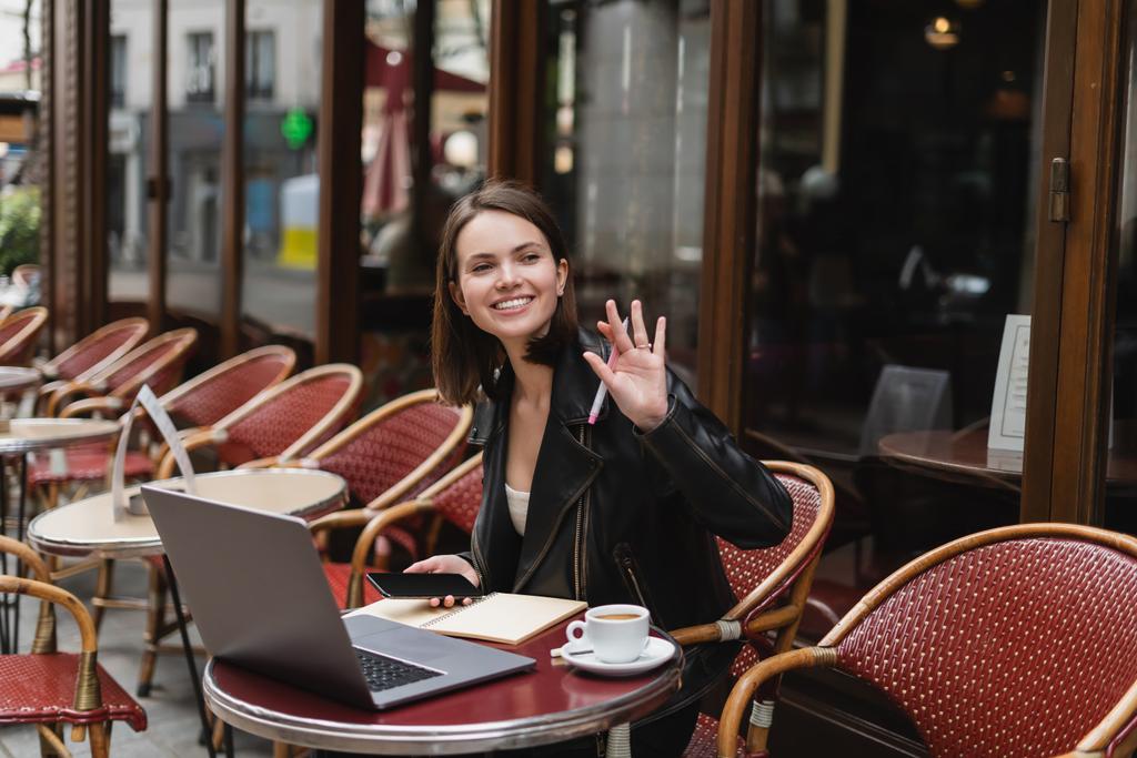 幸せな女性で黒のジャケットを保持スマートフォンと手を振って近くのノートパソコンとコーヒーカップでフランスの屋外カフェ  - 写真・画像