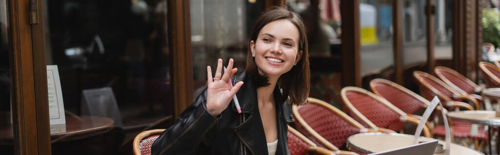 szczęśliwa kobieta w czarnej kurtce trzymając pióro i machając ręką w francuskiej kawiarni na świeżym powietrzu, baner - Zdjęcie, obraz