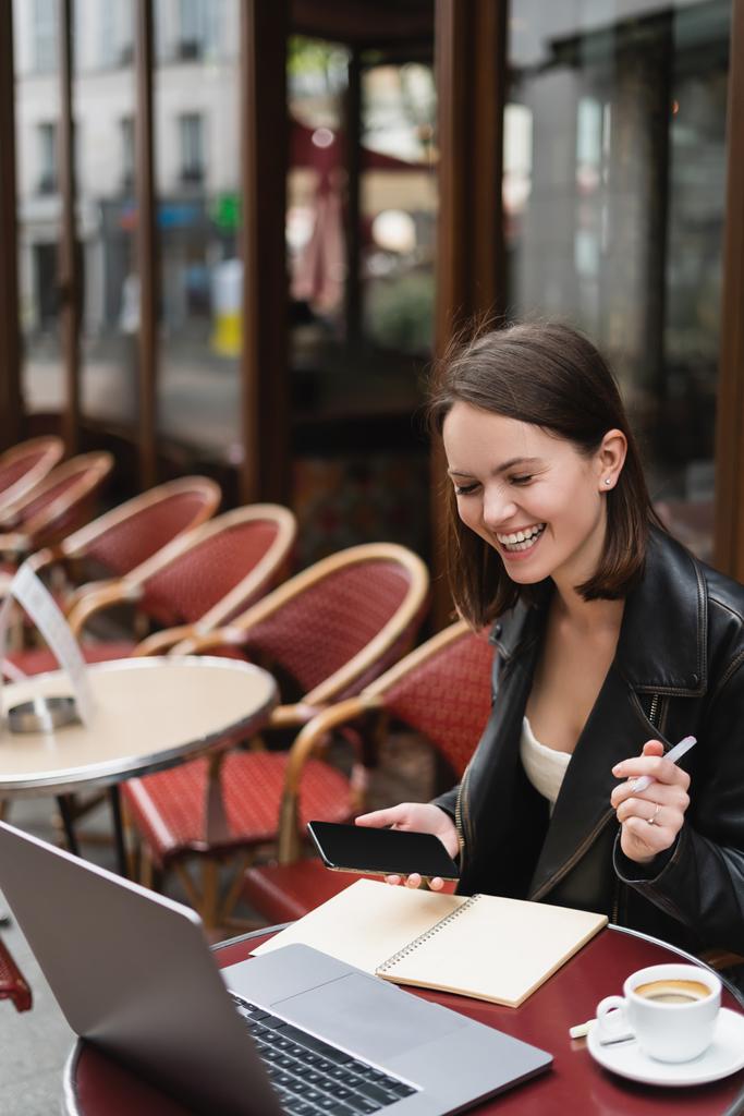 フランスの屋外カフェでスマートフォンやペンを持っている黒のジャケットで陽気なフリーランサー  - 写真・画像