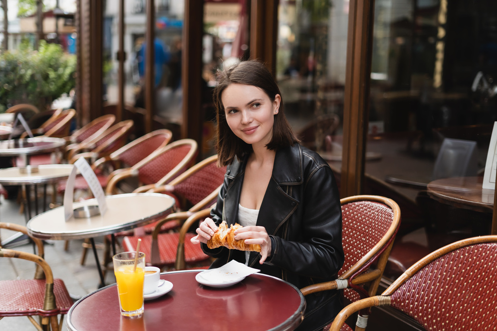 mujer alegre sosteniendo croissant cerca de taza de café y vaso de jugo de naranja en la cafetería al aire libre en París - Foto, imagen