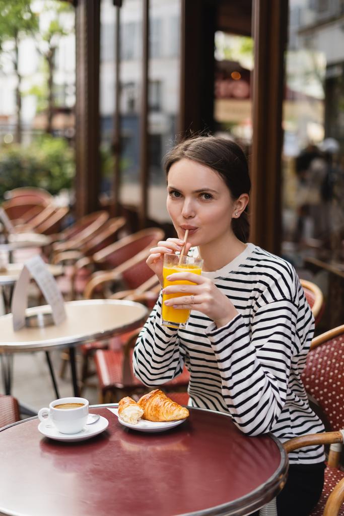 Nuori nainen raidallinen pitkähihainen paita tilalla lasi ja juo tuoretta appelsiinimehua ulkona kahvilassa Pariisissa - Valokuva, kuva