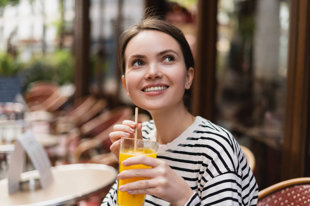 happy woman in striped long sleeve shirt holding glass of fresh orange juice in outdoor cafe in paris - Photo, Image