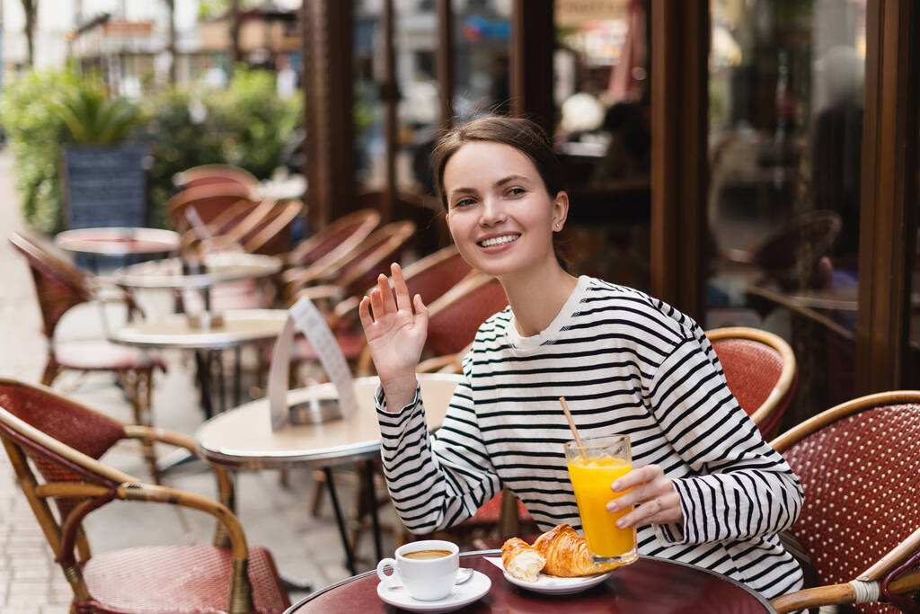 hymyilevä nainen raidallinen pitkähihainen paita tilalla lasi appelsiinimehua ja heiluttaa käsi ulkona kahvilassa Pariisissa - Valokuva, kuva