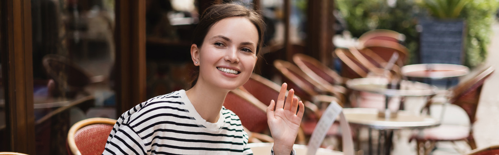 donna sorridente in camicia a righe che sventola mano in caffè all'aperto a Parigi, banner - Foto, immagini