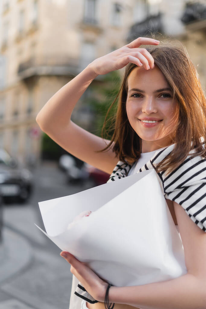 молодая женщина в стильном наряде регулирует волосы и держит букет завернутый в бумагу на улице в Париже  - Фото, изображение