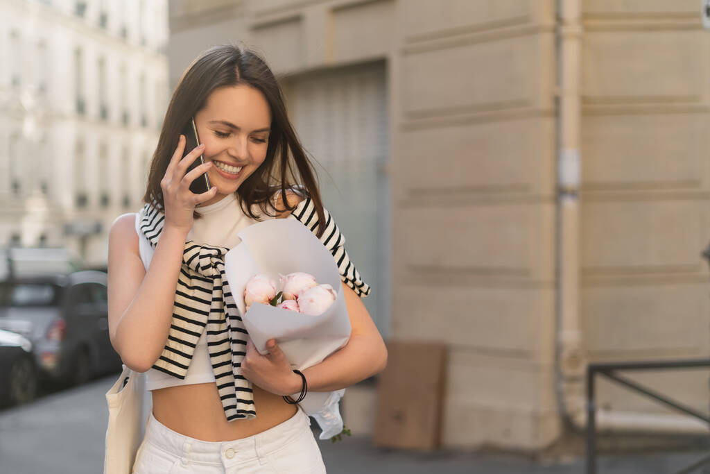 Moda kıyafetleri içinde mutlu bir kadın cep telefonuyla konuşuyor ve Paris 'teki şehir sokağında şakayık çiçekleri tutuyor.  - Fotoğraf, Görsel
