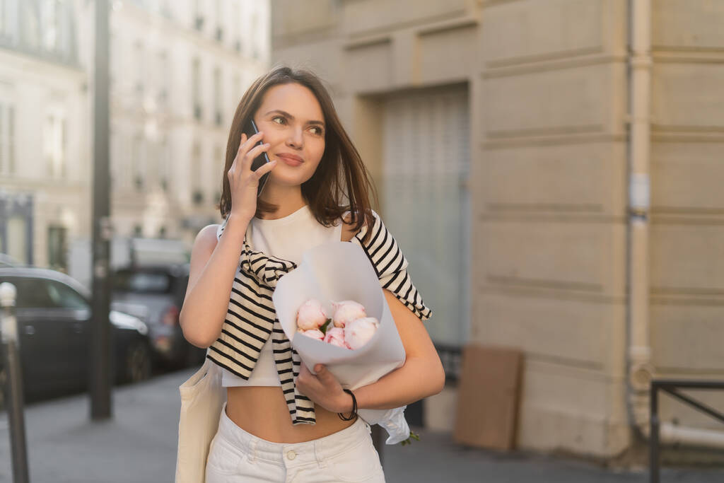 mujer sonriente hablando por teléfono móvil y sosteniendo ramo con peonías en la calle urbana en París  - Foto, imagen