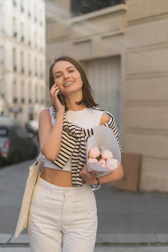 pleased woman talking on mobile phone and holding bouquet with peonies on urban street in paris  - Photo, Image