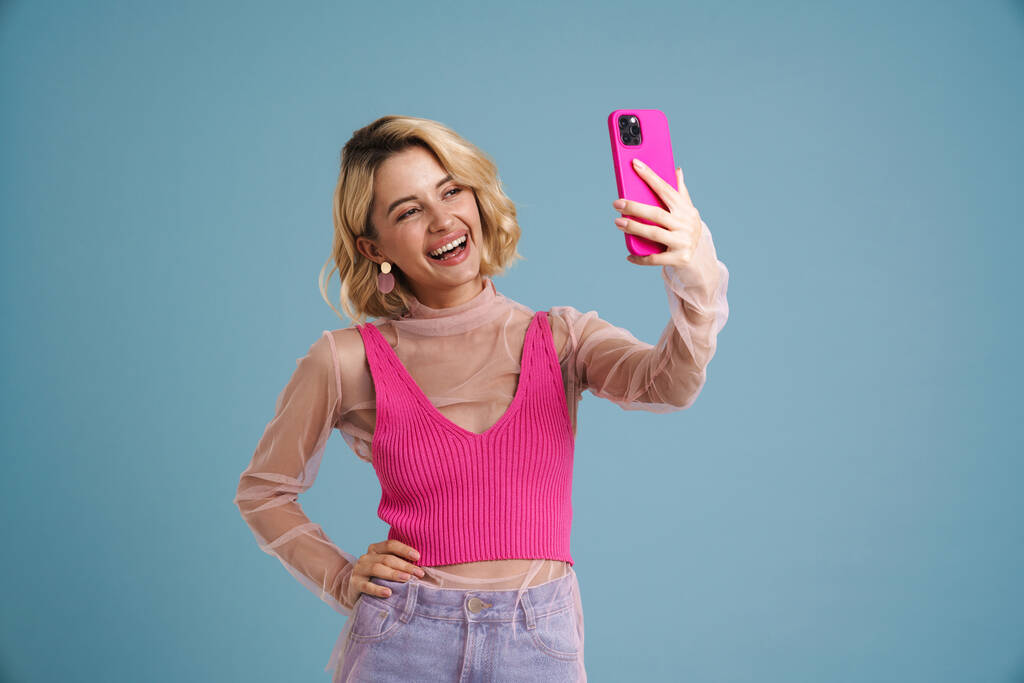 Giovane donna bianca sorridente mentre prende selfie sul cellulare isolato su parete blu - Foto, immagini