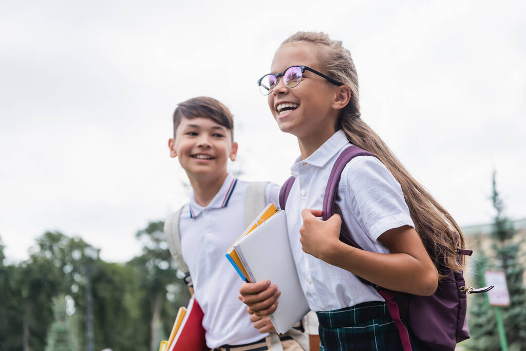 Pozitív iskolás gyerek hátizsákkal és notebookokkal sétál a homályos ázsiai barát közelében a szabadban  - Fotó, kép