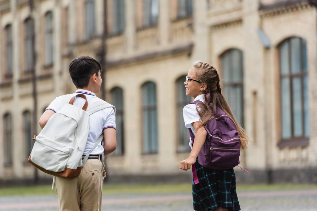 Θετική μαθήτρια με σακίδιο που μιλάει σε φίλο ενώ περπατάει κοντά σε θολό σχολείο  - Φωτογραφία, εικόνα