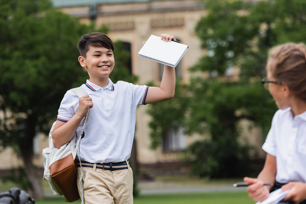 Азиатский школьник держит книгу рядом с размытым одноклассником в парке  - Фото, изображение