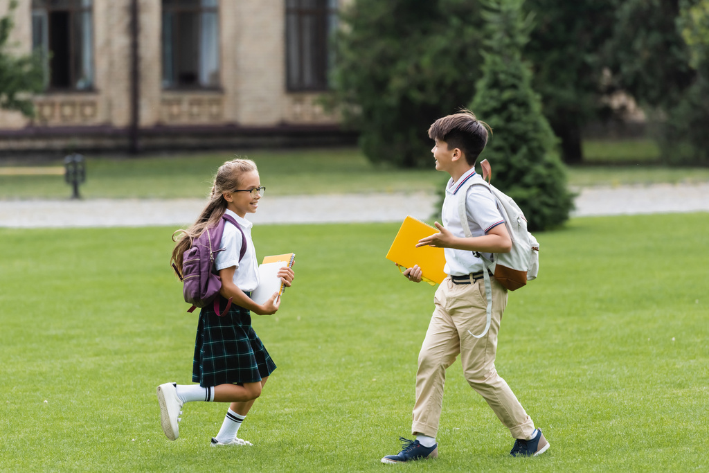 Uśmiechnięte wielonarodowe koleżanki z klasy z notatnikami biegającymi po trawniku w parku  - Zdjęcie, obraz